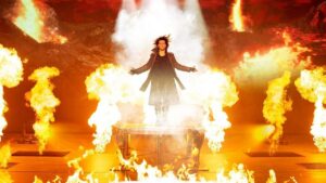 Man standing on a box with this hands out to his side with flames all around him